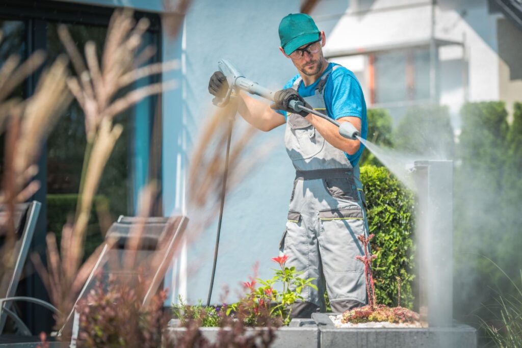 A professional pressure washing technician wearing safety gear and work overalls uses a high-powered washer to clean an outdoor surface near a residential building